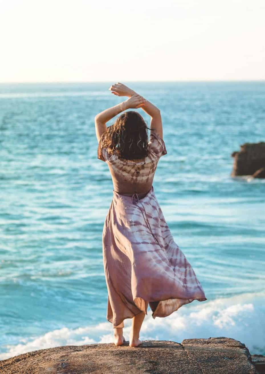 Frau in Kleid steht entspannt auf einem Felsen am Meer mit den Armen über dem Kopf